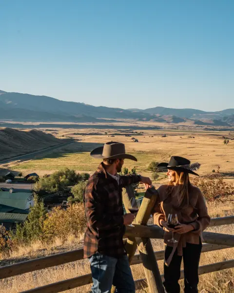 A couple wearing cowboy hats holding glasses of wine while standing outdoors overlooking a scenic valley.