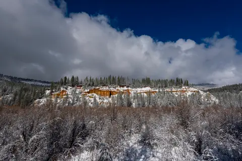 A snowy landscape features a rocky cliff face topped with trees under a partly cloudy blue sky.