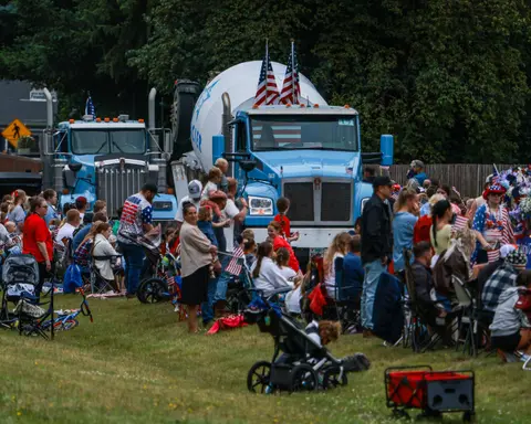 A crowd of people gathers along a street to watch a parade featuring large trucks.