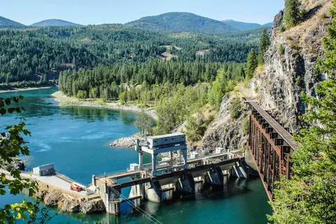 A dam on a river next to a railroad bridge and forested mountains.