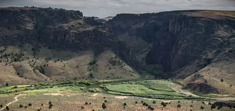 A river flows through a wide canyon with steep, rocky cliffs and green vegetation along its banks.