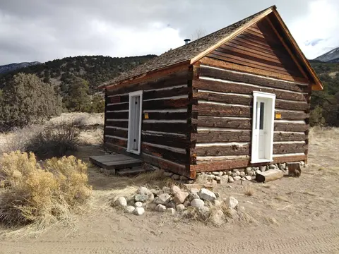 A historic log cabin stands in a dry, mountainous landscape.