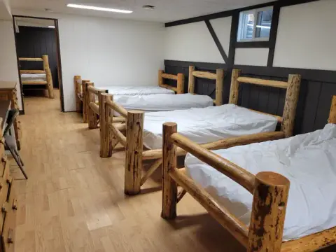 A dormitory-style room features multiple rustic log-style beds with white sheets.
