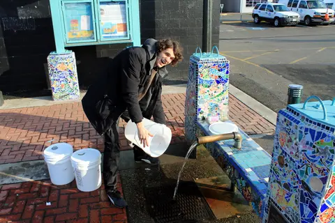 A smiling man fills white buckets from an outdoor artesian well structure.