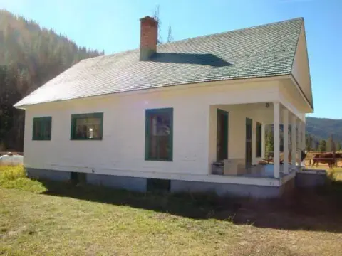A white historic house with a green roof and a front porch sits in a grassy field.