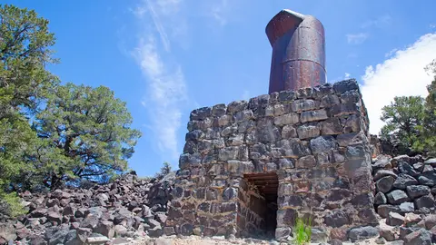 A stone and metal ruin stands among trees and rocks under a blue sky.