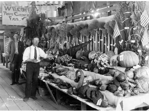 Two men stand beside a large agricultural produce exhibit decorated with flags.