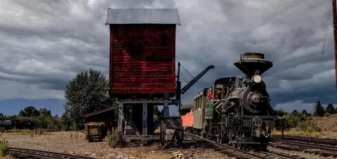 A historic steam train stopped by a red water tower under a cloudy sky.