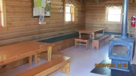 The interior of a log cabin features wooden tables, benches, and a stove.