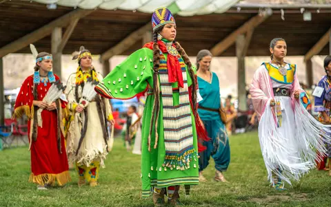 Several people wearing traditional indigenous regalia stand in a grassy area during a gathering.