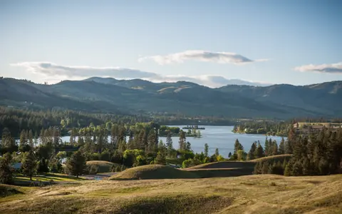 A scenic view of rolling hills, forests, and a body of water under a blue sky.