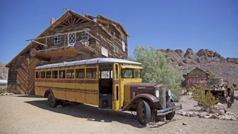 An old bus parked in front of a rustic wooden building in a desert landscape.
