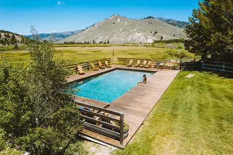 A person sits on the edge of an outdoor swimming pool with mountains in the background.