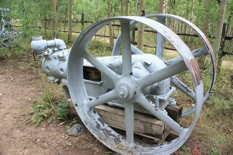 An old grey engine with large flywheels sits in a forest clearing.