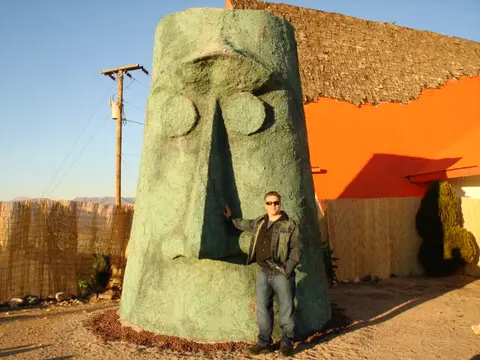 A man stands next to a large stone head statue outside an orange building.