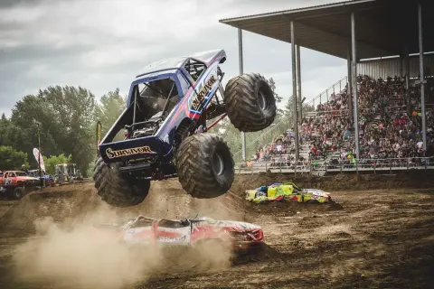 A monster truck jumps over crushed cars in front of a grandstand filled with spectators.