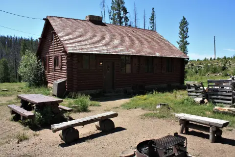 A rustic log cabin stands in a clearing with a picnic table and a fire ring.