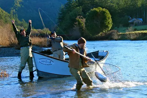 Three men fishing in a river, with one netting a fish, another cheering, and another holding a fishing rod in a boat.