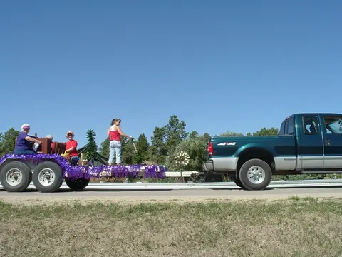 A green pickup truck pulls a decorated trailer carrying people outdoors.