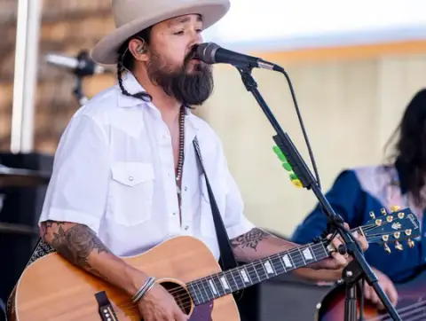 A man wearing a hat and white shirt plays acoustic guitar and sings into a microphone.