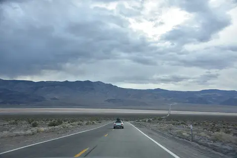 A car travels down a long road through a desert landscape under a cloudy sky.