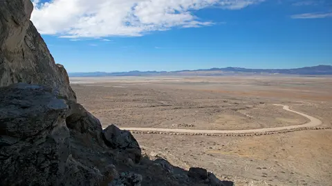 A vast desert landscape viewed from an elevated rocky area with a winding road below.