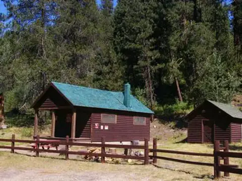 A wooden cabin sits surrounded by pine trees with a fence in the foreground.