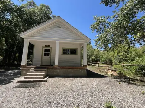 A small white building with a covered porch stands outdoors amidst trees and a fence.