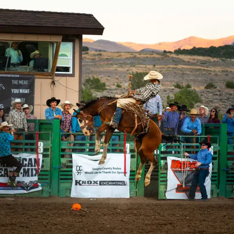 A cowboy rides a bucking horse at a rodeo arena.