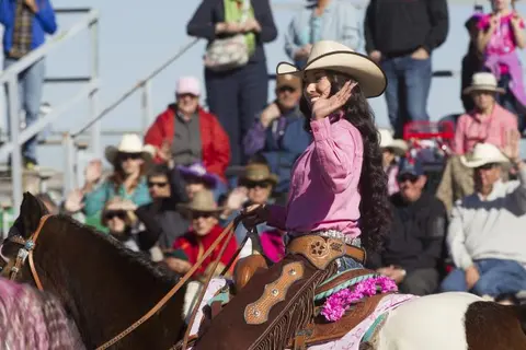 A woman in a cowboy hat and pink shirt rides a horse while waving to a crowd.