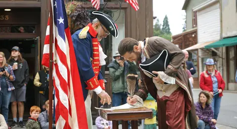 Two men in historical colonial attire participate in an outdoor ceremony near an American flag.