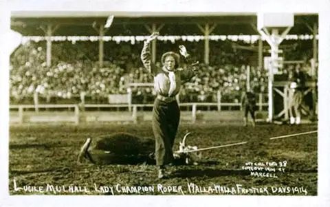 A historic black and white photograph showing a roper standing in an arena in front of a crowd.
