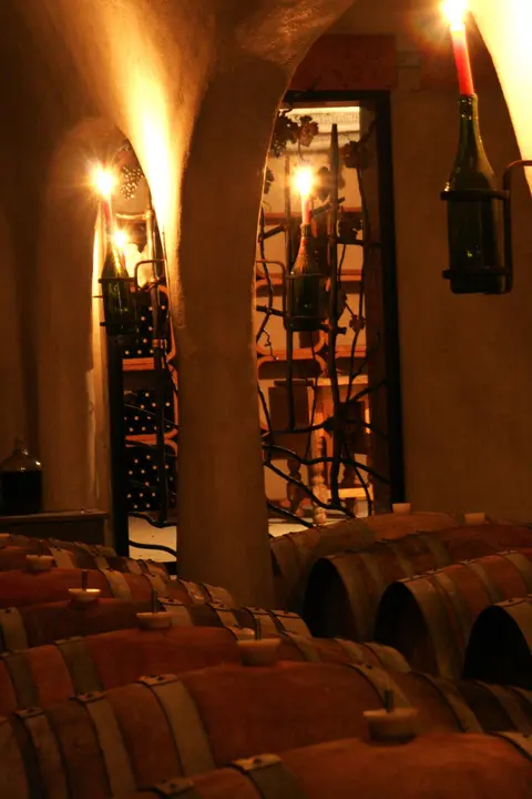 Rows of wooden wine barrels rest inside a dimly lit cellar illuminated by candles in bottles.