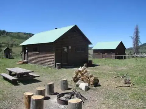 A rustic log cabin in a grassy clearing with a fire pit and picnic table in the foreground.