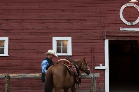 A cowboy stands beside a saddled horse in front of a red barn.