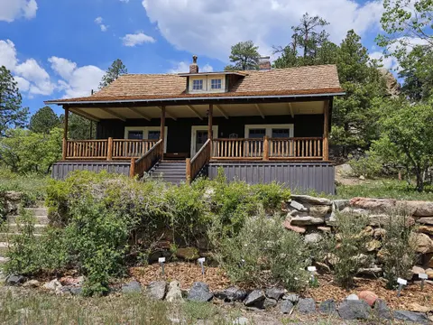 A dark wooden cabin with a covered porch stands behind a stone retaining wall and greenery under a partly cloudy sky.