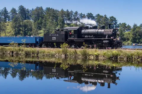 A steam train travels along a body of water, reflecting on the surface.