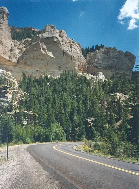A winding asphalt road curves past a steep forested hillside with rock cliffs above.