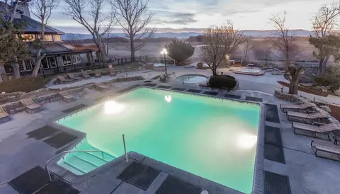 A lighted outdoor swimming pool and hot tubs are surrounded by lounge chairs and buildings at dusk.