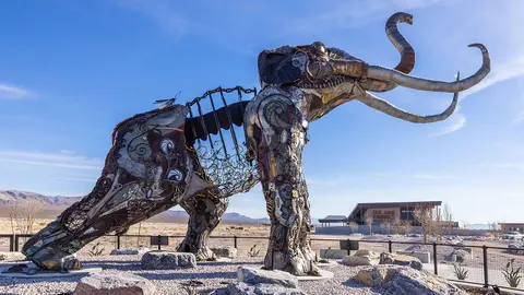 A large sculpture of a mammoth made of scrap metal stands outdoors against a blue sky.
