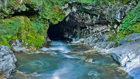 A river flows out of a dark cave surrounded by mossy rocks and vegetation.