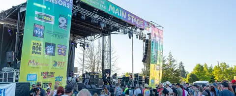A band performs on an outdoor stage at a fair while a crowd watches.
