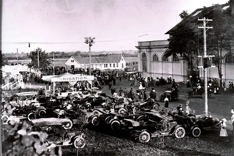 Historic black and white photo of a large gathering with many parked early automobiles.