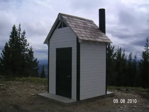 A small white outhouse with a dark roof and chimney sits on a mountain ridge.