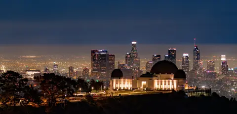A large domed observatory building is lit up at night with a city skyline in the background.