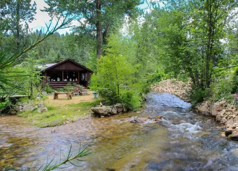 A rustic wooden cabin sits beside a rushing stream in a forested valley.