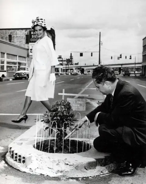 A historic black and white photo shows a man tending to a small fenced park in the middle of a street while a woman walks nearby.