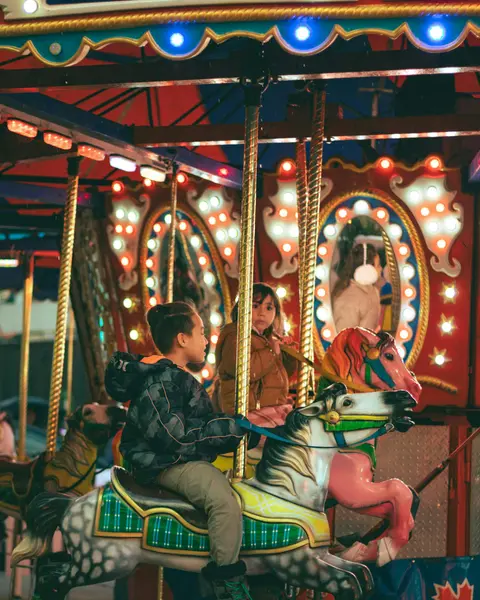 Two children riding horses on a lit carousel at night.
