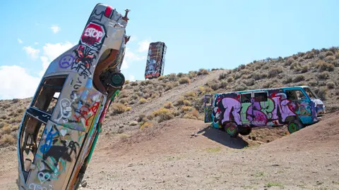 A car and vans covered in graffiti stand upright and on their sides in a desert landscape.