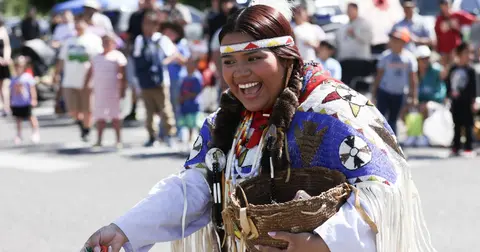 A smiling person in traditional regalia holds a woven basket while participating in an outdoor event.